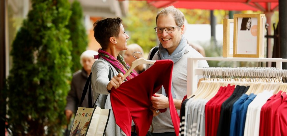 Ein Pärchen mittleren Alters kauft Kleidung. Die Dame zeigt Ihrem Mann ein rotes Langarmshirt.