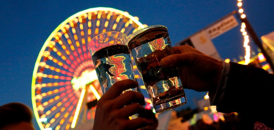 Vor einem beleuchteten Riesenrad, es ist Nacht,  fällt der Blick auf zwei gefüllte Dubbegläser mit Weinschorle, die gerade mit einander anstoßen.