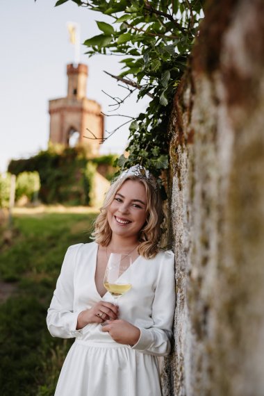 Die Dürkheimer Weinprinzessin Denise Stripf angelehnt an eine Sandsteinmauer. Im Hintergrund ist der Bad Dürkheimer Flaggenturm zu sehen.