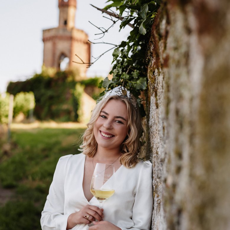 Die Dürkheimer Weinprinzessin Denise Stripf angelehnt an eine Sandsteinmauer. Im Hintergrund ist der Bad Dürkheimer Flaggenturm zu sehen.