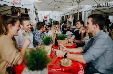 Verschiedene Personen sitzen an einem Tisch im Weindorf auf dem Wurstmarkt. Sie trinken Wein, unterhalten sich und lachen.